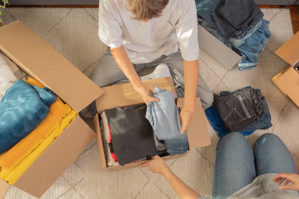 Woman and child sorting clothes and packing into cardboard box. Donations for charity, help low income families, declutter home, sell online, moving moving into new home, recycling, sustainable living stock photo