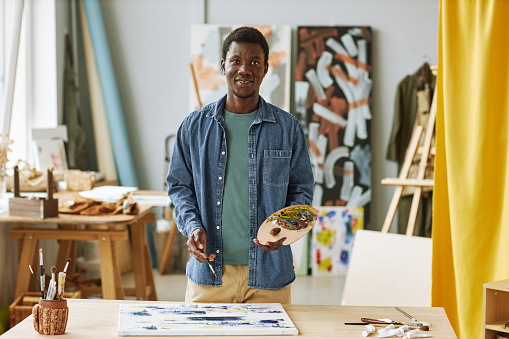 Young successful African American male artist in casualwear holding wooden color palette with acrylic paints while creating artwork