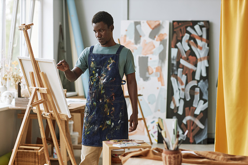Young serious African American man in denim apron standing in front of canvas on easel and creating new painting at lesiure