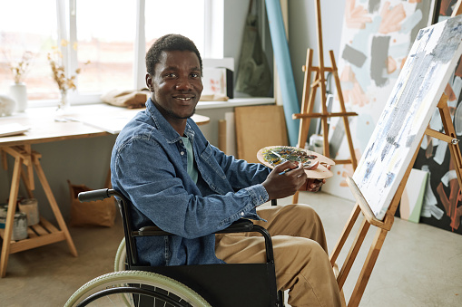 Young successful painter with disability sitting in wheelchair in front of camera and looking at you while working over new painting