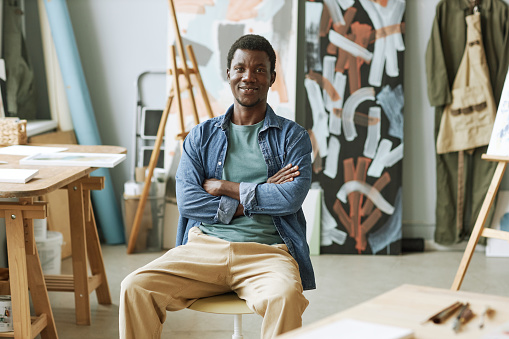 Happy young African American man in casualwear sitting in front of camera against easels, wooden table and abstract paintings in studio
