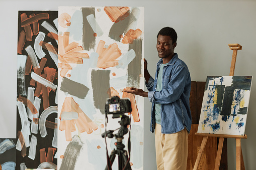 Young smiling blogger or painting coach presenting new abstract painting created by him while standing in front of video camera