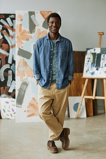 Happy young creative craftsman in casual attire standing in front of camera in studio of arts against collection of his paintings