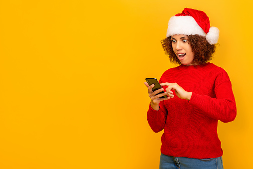 Pleasantly surprised African American woman reading message on mobile phone about big christmas sales or winning lottery standing on orange background. Good news, discounts, online shopping concept