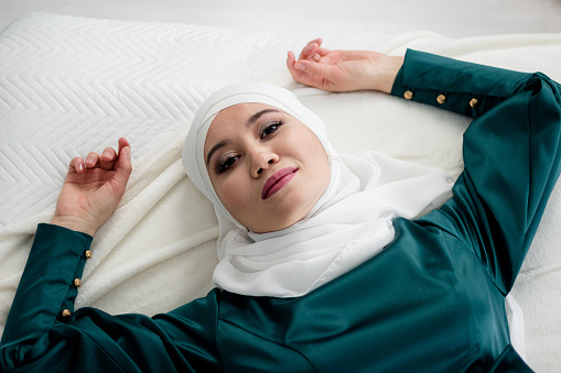 A Muslim woman in a beautiful green dress in a white headscarf lies on the bed, close-up. Relaxation.
