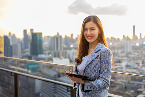 Young Asian business woman in formal suit with digital tablet standing outside the skyscraper building for real estate, housing and urban development