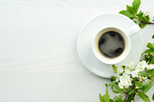 Coffee and flowers. Black hot coffee in a white coffee cup and a blooming apple tree branch on a white wooden background or table