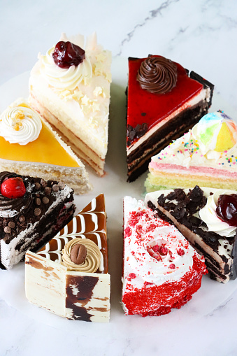 Stock photo showing close-up, elevated view of slices of chocolate and cherry cake, Black Forest gateau, red velvet, rainbow cake, coffee and marbled chocolate gateau displayed on a circle of greaseproof parchment paper against a marble effect background.