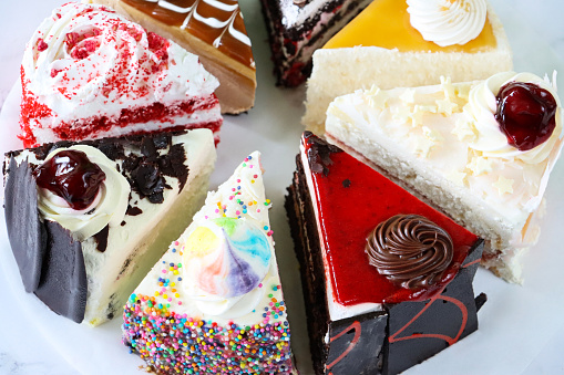 Stock photo showing close-up, elevated view of slices of chocolate and cherry cake, Black Forest gateau, red velvet, rainbow cake, coffee and marbled chocolate gateau displayed on a circle of greaseproof parchment paper against a marble effect background.