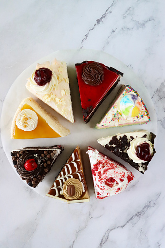 Stock photo showing elevated view of slices of chocolate and cherry cake, Black Forest gateau, red velvet, rainbow cake, coffee and marbled chocolate gateau displayed on a circle of greaseproof parchment paper against a marble effect background.