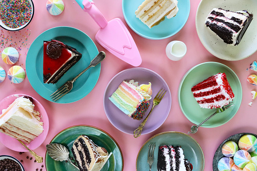 Stock photo showing close-up, elevated view of multicoloured plates containing slices of chocolate and cherry cake, Black Forest gateau, red velvet, rainbow cake, coffee and marbled chocolate gateau displayed on a pink background.