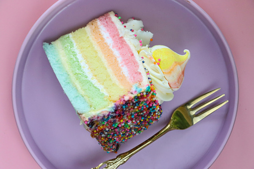 Stock photo showing close-up, elevated view of a slice of rainbow cake displayed on a plate against a pink background.