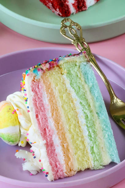 istock Close-up image of rainbow cake slice, five rainbow sponge layers separated by vanilla buttercream on purple plate with fork, piped icing rosette, rainbow meringue kiss, red velvet cake on green plate, pink background, focus on foreground 1442893195