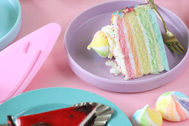 istock Close-up image of rainbow cake slice, five rainbow sponge layers separated by vanilla buttercream on purple plate with fork, piped icing rosette, rainbow meringue kiss, pink background 1442893197