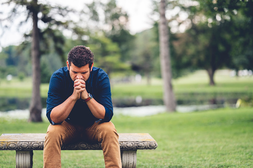 A young Caucasian man praying while sitting on a bench