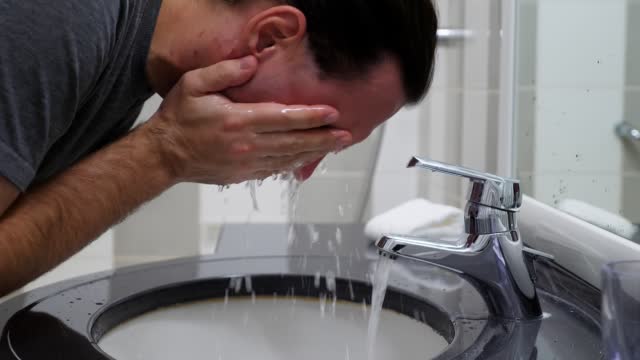 Man pours water into his palms and rinses his face, standing leaning over sink