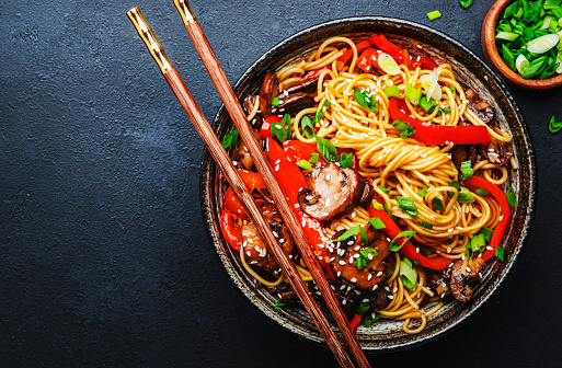 istock Vegan stir fry egg noodles with vegetables, paprika, mushrooms, chives and sesame seeds in bowl. Asian cuisine dish. Black table background, top view 1442924073