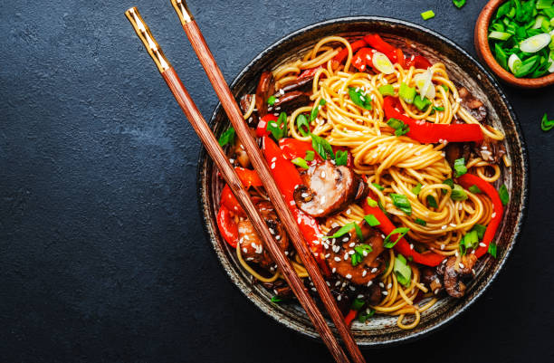 Vegan stir fry egg noodles with vegetables, paprika, mushrooms, chives and sesame seeds in bowl. Asian cuisine dish. Black table background, top view stock photo