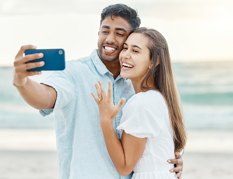 Love, beach and engaged couple taking a selfie on a phone while on a romantic summer vacation. Happy woman showing off her wedding ring while taking a picture with her husband on honeymoon holiday.