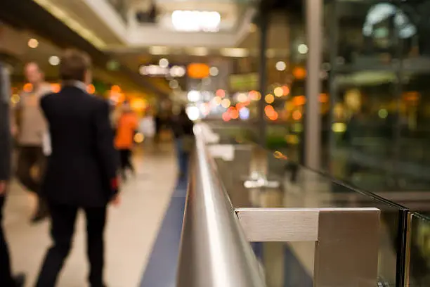 In a Shopping mall or railstation. Focus on Handrail in foreground. Background and peopleout of focus