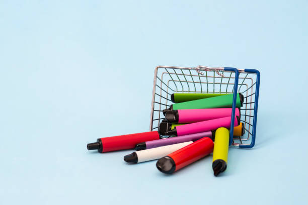 um conjunto de bastões multicoloridos - dispositivos vaping descartáveis, cigarros eletrônicos em uma cesta de compras em um fundo azul. - tobacco wagon - fotografias e filmes do acervo