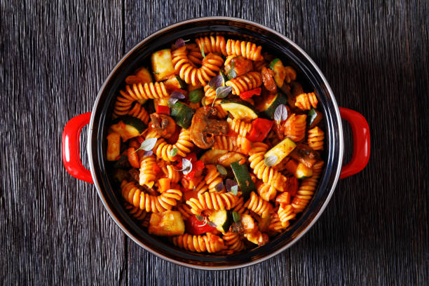 One Pot Vegetable Pasta of rotini, zucchini, capsicum, mushrooms smothered in a garlic herb tomato sauce in red pot on dark wood table , flat lay, close-up