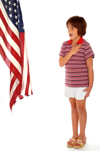 Young elementary girl dressed in American colors saying the Pledge of Allegiance towards the American flag.  Isolated on white.