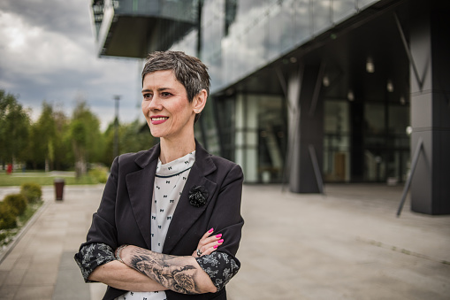 Portrait of a modern businesswoman with short hair and tattoos on her arm, standing in front of a modern office building