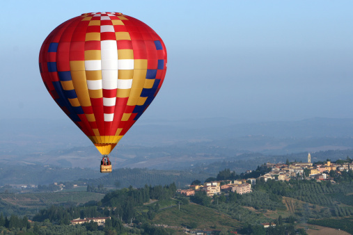 Panoramic photograph of a brightly colored hot air balloon flying over a small village in the Tuscany region of Italy.