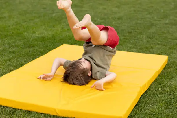 5 year old cheerful white boy doing gymnastic exercises on a soft mat in the yard of his house