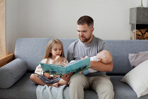 Young single dad with two children sitting on couch at home, reading book to his daughter and holding newborn baby in his arms. Family time