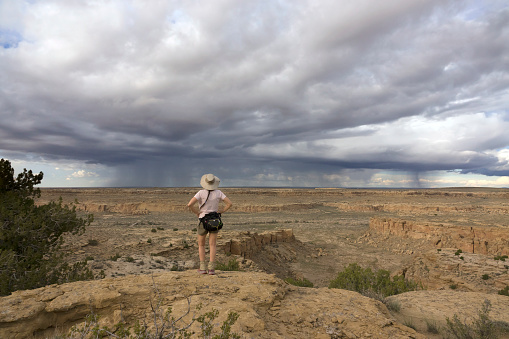 Hiking the South Mesa Trail in Chaco Culture National Historical Park with falling rain and thunderheads overhead, a woman walks past a distant view of Pueblo Bonito and Pueblo Alto in the distance.