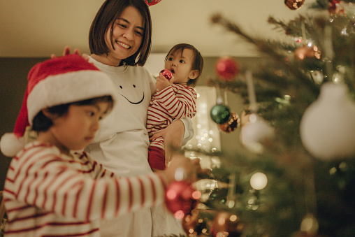 Mother helping her cute boy in decorating christmas tree. A boy in a Santa hat is sitting on the floor and decorating a christmas tree and smiling.