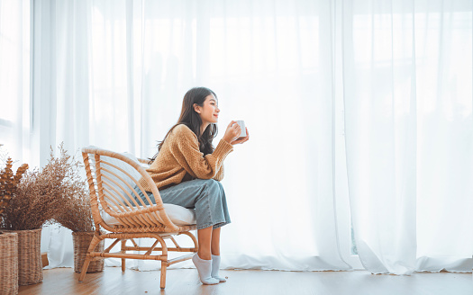 Happy asian woman relaxing drinking hot coffee or tea in holiday morning vacation on armchair at home, Cosy scene, Smiling pretty woman drinking hot tea in autumn winter. copy space.