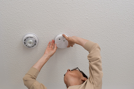 Middle aged woman reaching up for smoke alarm on ceiling at home, changing battery, installing or regular maintenance concept
