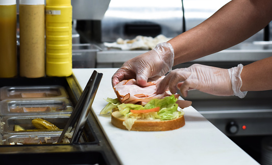 Kitchen employee preparing turkey deli meat sandwich with lettuce for customer food order using protective gloves