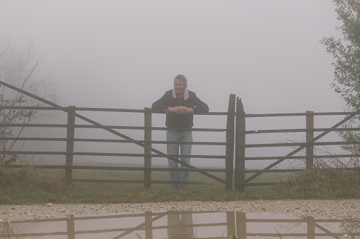 Farmer leaning on his wooden farm fence gate on foggy autumn day