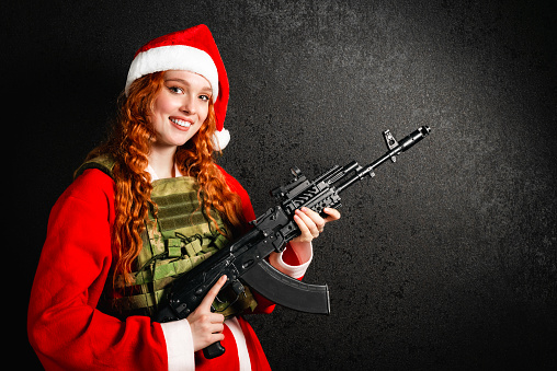Woman in a Santa Claus costume and a red hat holds an assault rifle in her hands. Portrait of a girl in an army bulletproof vest on a black dark background. Concept of military Christmas, New Year.