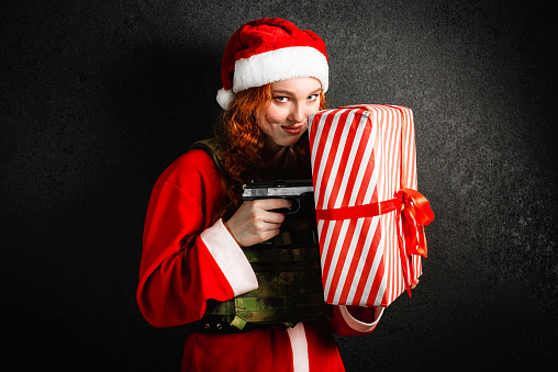 A woman in a Santa Claus costume and a red hat is holding a gun. Portrait of a girl in an army bulletproof vest with a gift box on a black dark background. Concept of military Christmas, New Year.