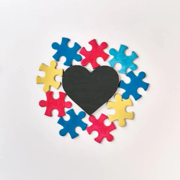 Autism campaign symbols. Colorful puzzle pieces around blackboard stone heart with copy-space for your text, lettering. Flat lay, top view, square composition on off white background.