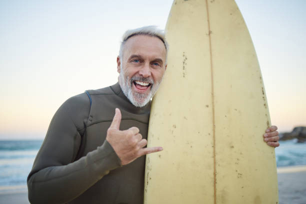 senior man, with surfboard and at beach smile, happy and at sunset with swimwear on a holiday and vacation. portrait, shaka and older male surfer, hand sign and at seaside surfing while on a trip. - senior adult human face male action imagens e fotografias de stock