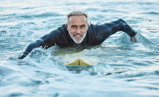 Man is at the beach during summer, in the ocean and surfing with his surfboard in a portrait. Mature, smile and surf in a wet suit for sport, fitness and health and happy in the sea water.