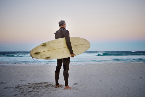 Surfer, surfboard and man on beach looking at sea waves in during sunset during summer vacation in Hawaii. Professional male athlete rest after training or practice surfing sport outdoor at the ocean