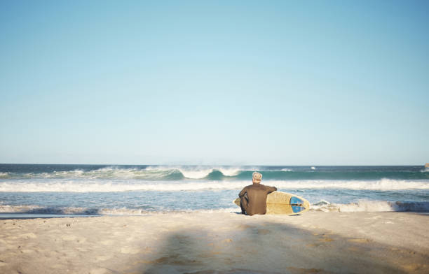 surf, mer et sports avec un homme sur la plage pour le surf, le fitness ou l’entraînement avec vue sur l’horizon et maquette du ciel. entraînement, exercice et santé avec un surfeur assis sur le sable avec sa planche de surf - senior adult outdoors wellbeing sky photos et images de collection