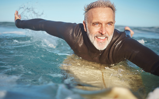 Beach, water and old man surfer swimming on a summer holiday vacation in retirement with freedom in Bali. Smile, ocean and senior surfing or body boarding enjoying a healthy exercise on sea of Island