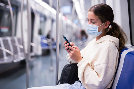 Active girl in a protective mask, riding on a subway train during a pandemic, corresponds with friends in a mobile 
phone