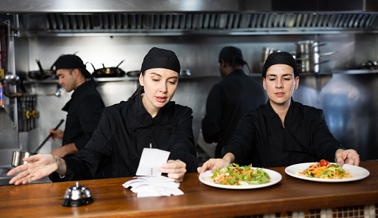 Portrait of two women chefs working in modern open kitchen, checking orders and giving out ready meals