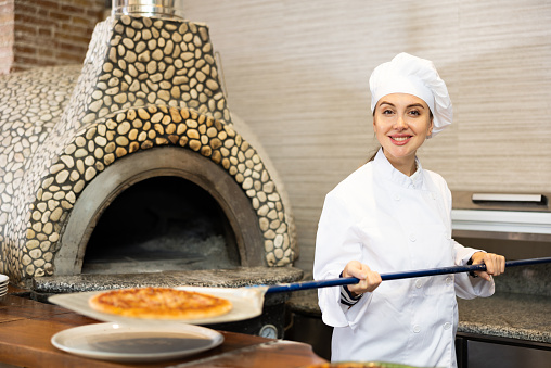Portrait of confident young woman chef working in restaurant kitchen, getting ready pizza out of oven