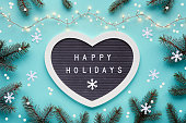 Text Happy Holidays on felt heart shape letter board, letterboard. Winter Christmas flat lay background with fir twigs, snowflakes, garland with Xmas lights. Monochromatic blue mint background.
