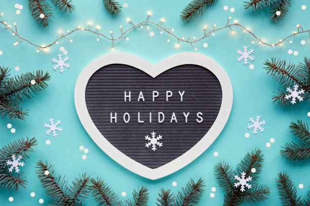 Text Happy Holidays on felt heart shape letter board, letterboard. Winter Christmas flat lay background with fir twigs, snowflakes, garland with Xmas lights. Monochromatic blue mint background. stock photo